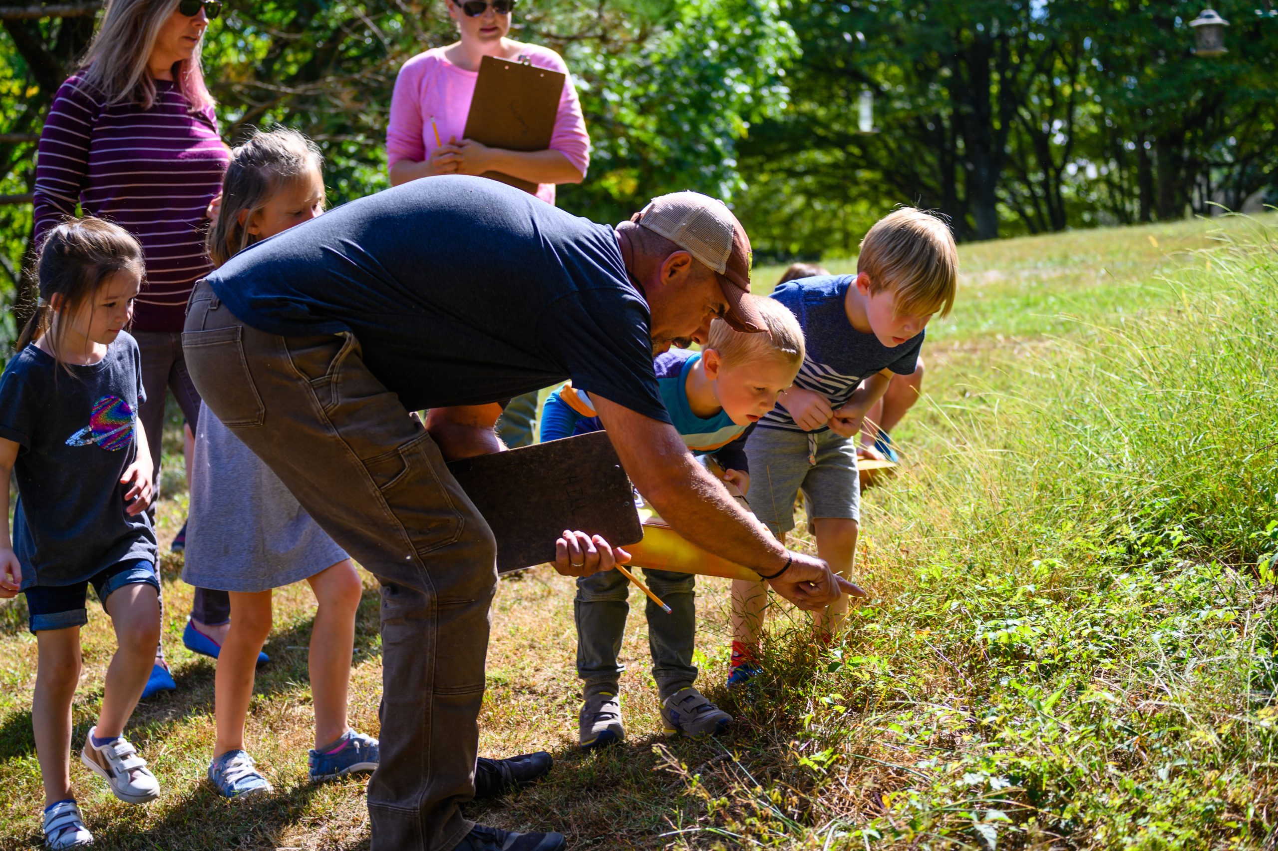 kids learning outside leaning over to look in grass at what teacher points